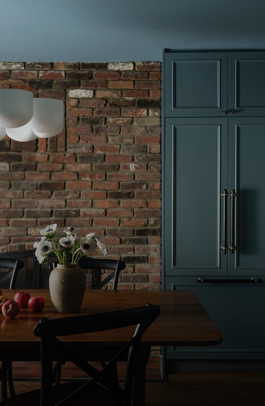Dining area with an exposed red-brick wall, blue-grey cabinetry and a wood table with black cross-back chairs