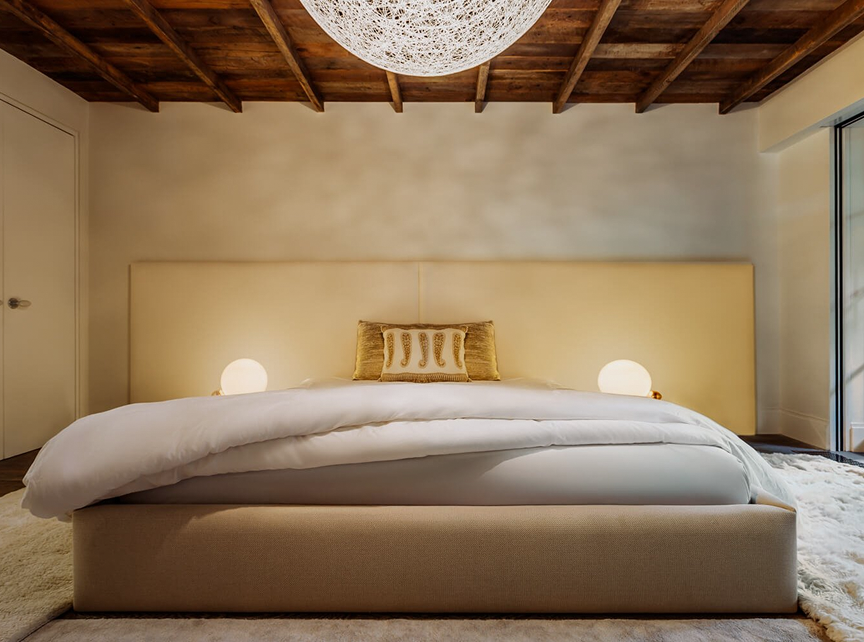 Bedroom with an upholstered cream bed and a woven pendant light beneath an exposed wood-beam ceiling