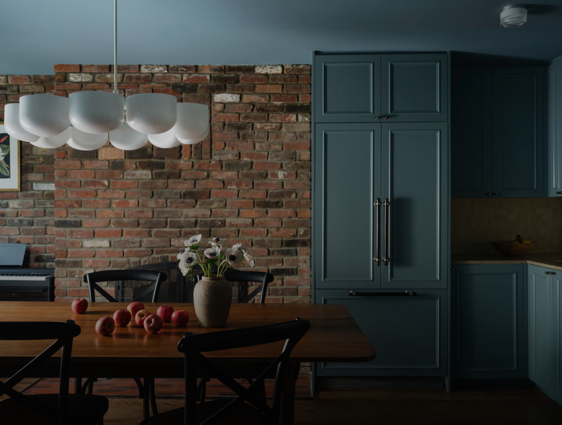 Dining area with an exposed red-brick wall, blue-grey cabinetry and a wood table with black cross-back chairs