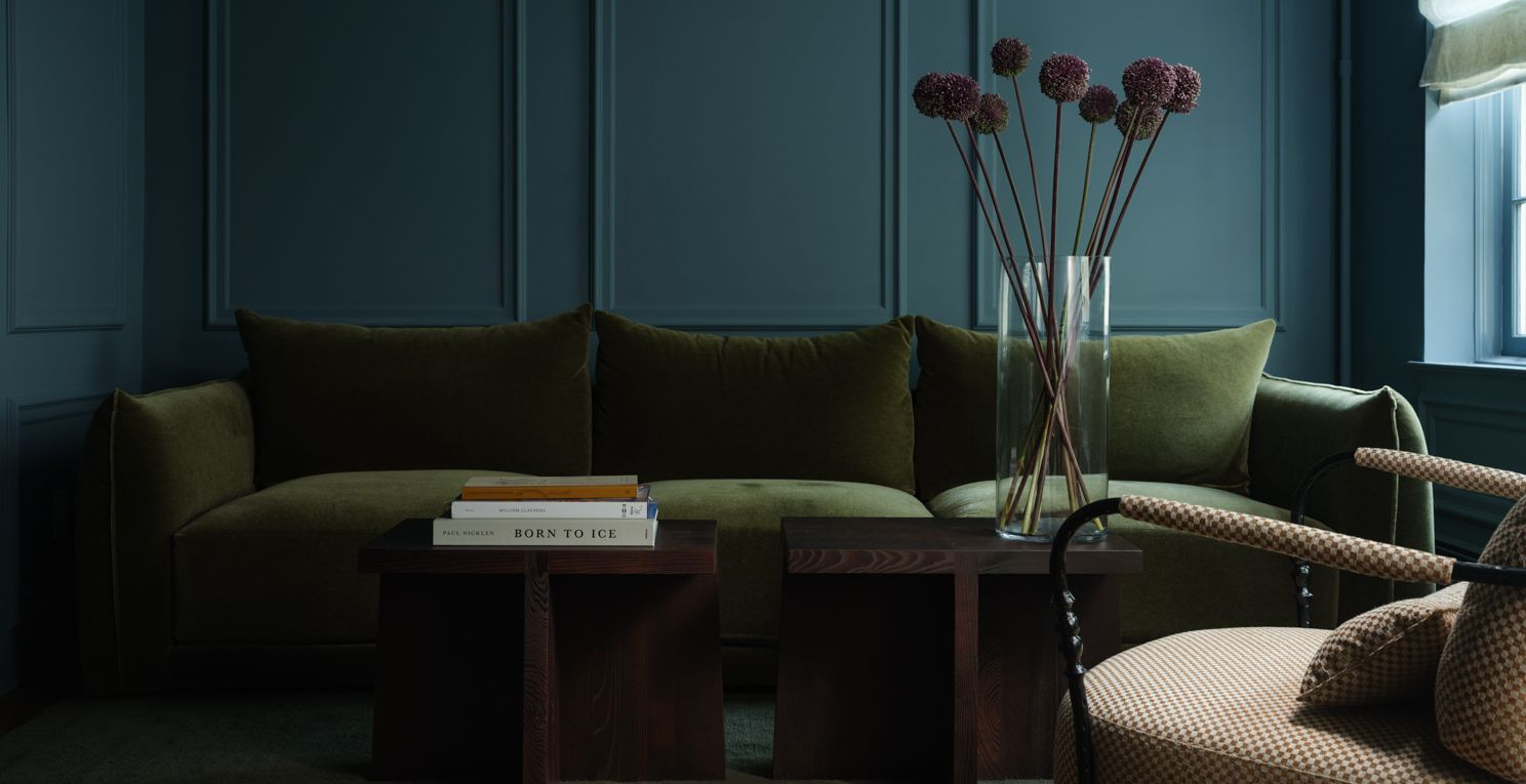 Sitting room with an olive-green velvet sofa against deep blue-green panelled walls and a vase of allium flowers