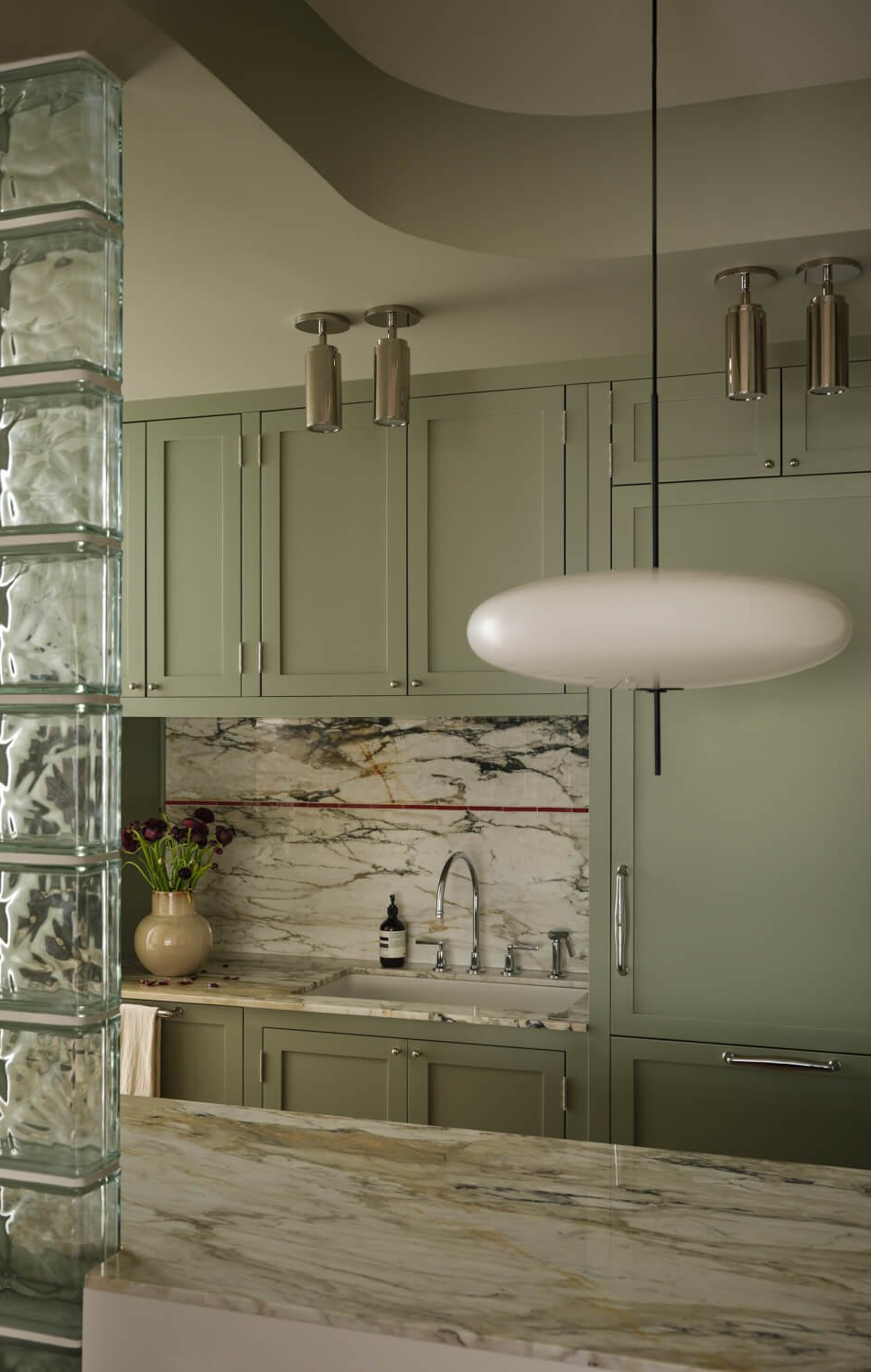 Sage-green kitchen with a veined-marble backsplash and island beside a glass-block wall