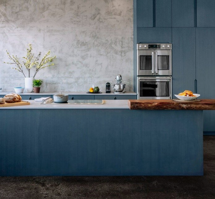 Blue kitchen with a concrete wall, double wall ovens and a live-edge counter